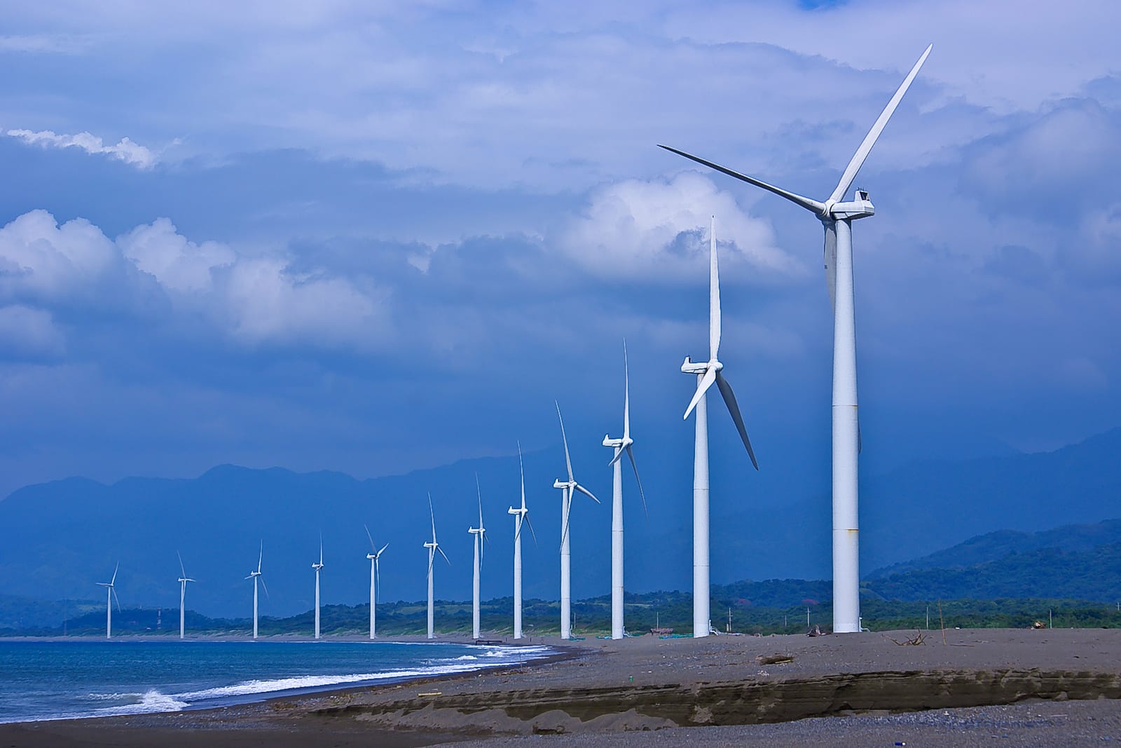 A long row of white wind turbines along a dark-sand beach, with surf on the left and blue mountains under a cloudy sky behind.