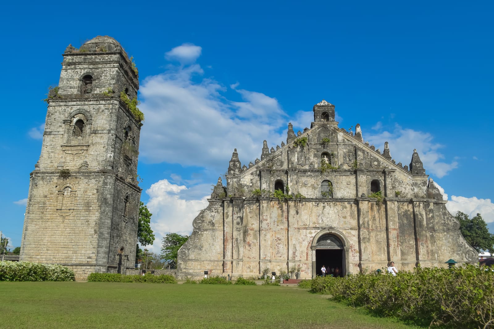 A massive old church of weathered coral stone and brick with huge buttresses, beside a separate square bell tower, on a lawn under a blue sky.