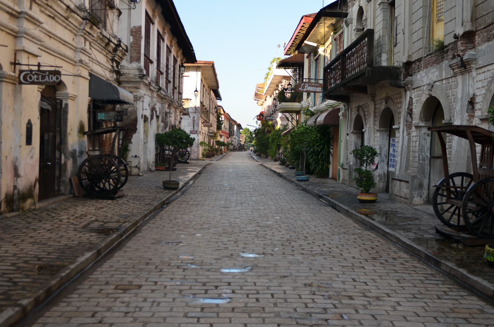 An empty cobblestone street between old two-storey stone houses with wooden balconies and capiz windows, cart wheels by the doors, in soft morning light.