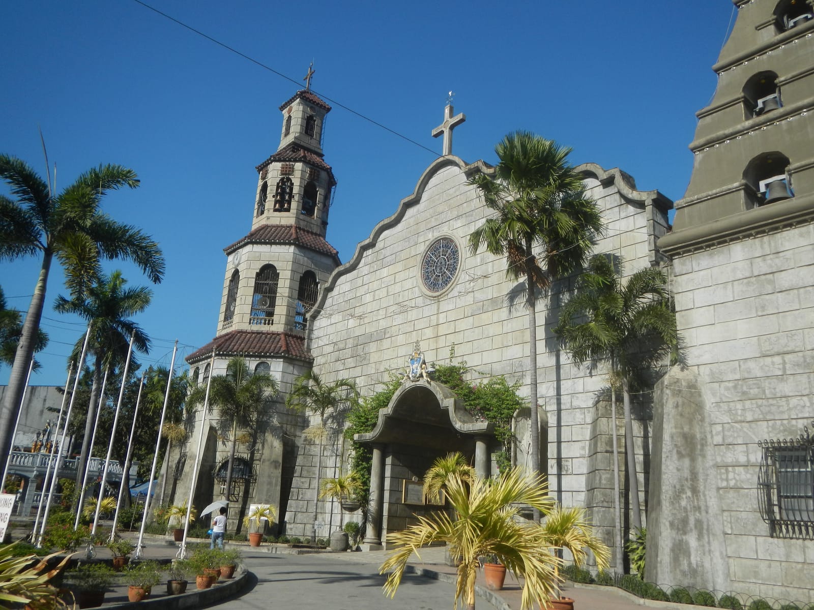 A grey stone church with a round window and a stepped gable, a tall tiered bell tower beside it and palms before it, under a deep blue sky.