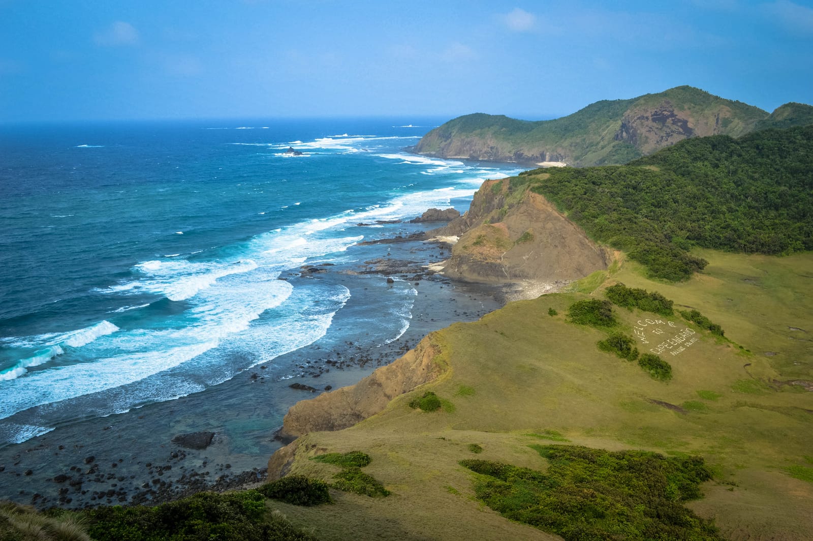 Grassy headlands and dark rocky shore along a deep blue sea with white surf, rising to a forested hill.
