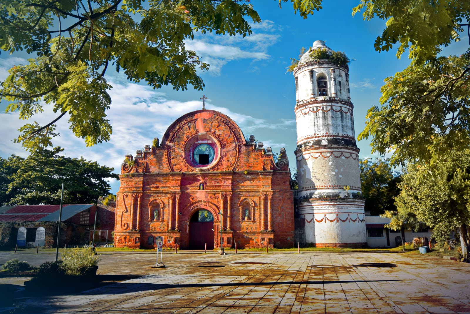 A red-brick baroque church with an ornate carved facade and a round window, beside a tall white cylindrical bell tower, framed by trees under a blue sky.