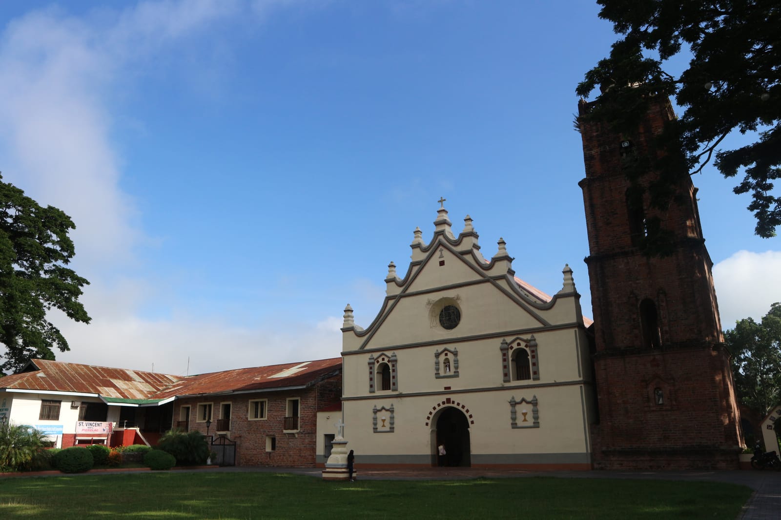 A cream church facade with a scrolled, pinnacled gable and a round window, beside a tall red-brick bell tower, behind a lawn under a blue sky.