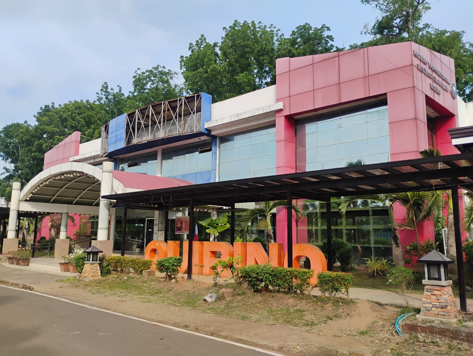 A pink-and-glass public building behind a covered walk, with large orange letters spelling “QUIRINO” among shrubs in front, under a pale sky.