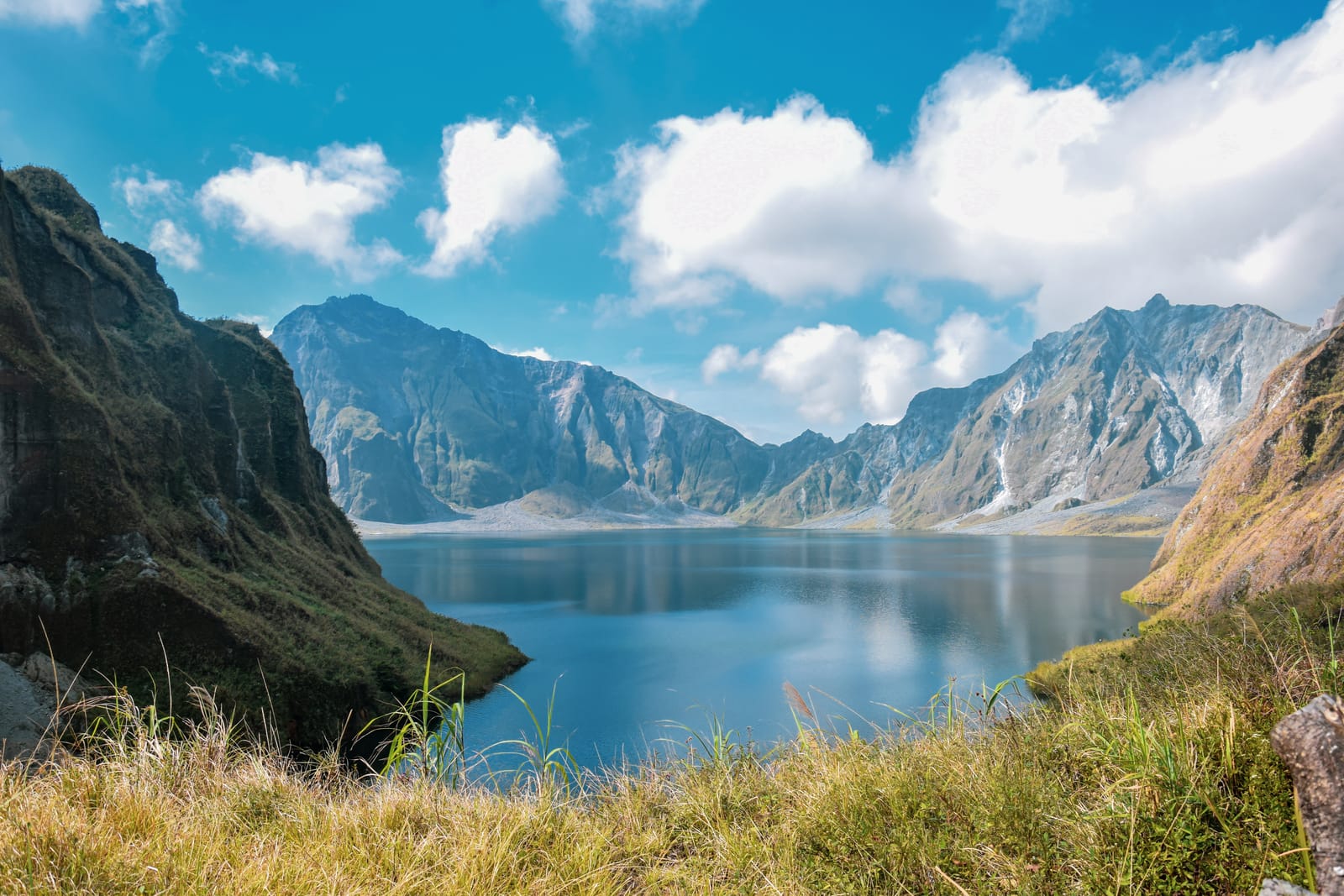 A blue crater lake ringed by steep grey and green cliffs, under a blue sky with white clouds, with tall grass in the foreground.