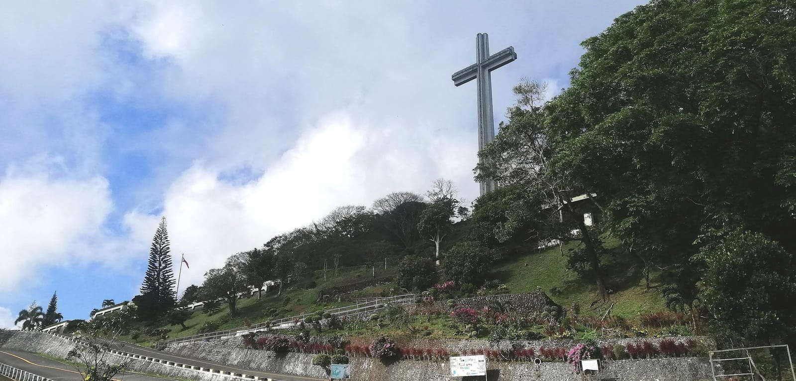 A towering concrete cross on a green hilltop above a landscaped slope of flower beds, under a blue sky with white clouds.