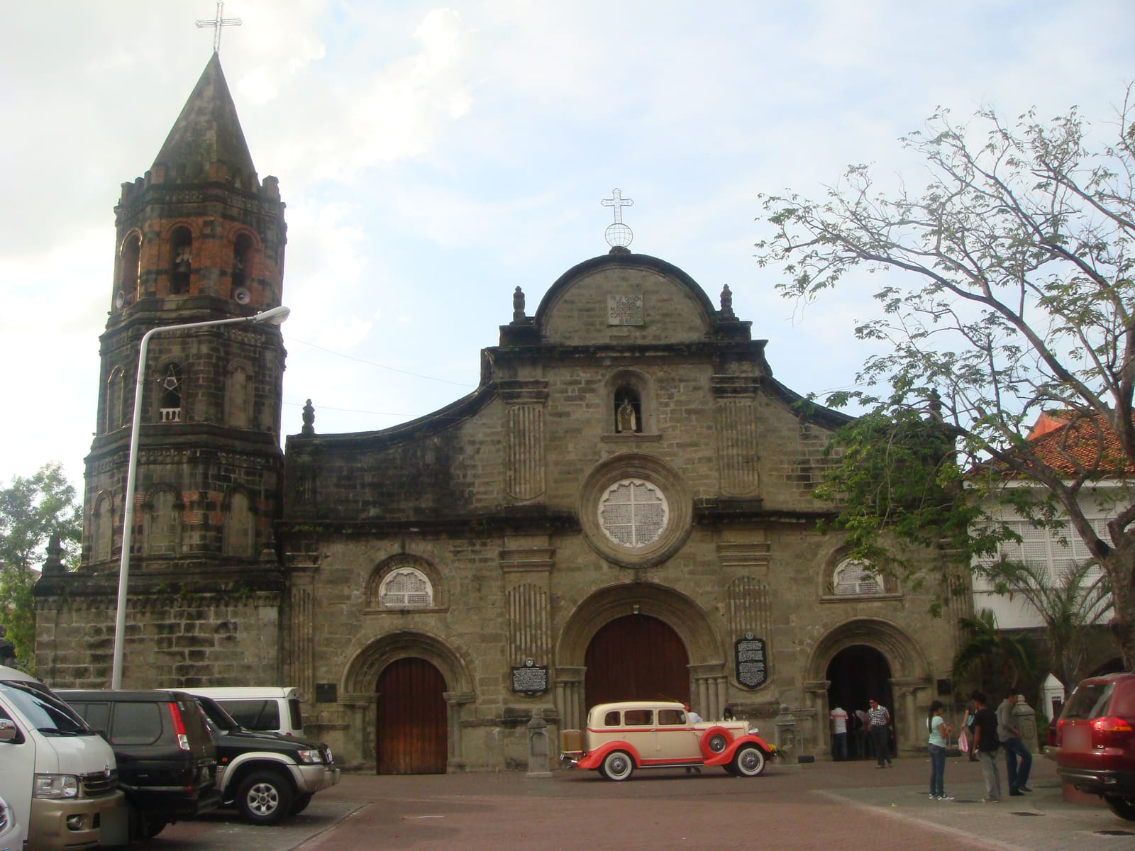 A weathered stone church with a round window and arched doors beside a tall bell tower, a vintage cream-and-red car parked before it and other cars to the left.