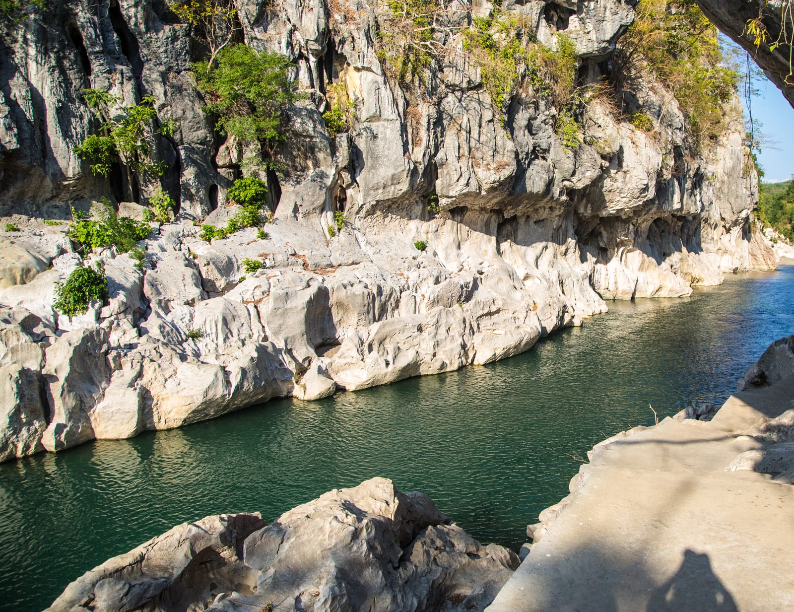 A green river running between towering white limestone walls, seen from a rock ledge in the shade of an overhang.