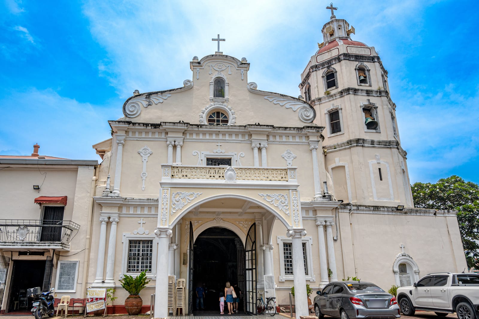 A cream-white church with a scrolled facade, an arched porch and a tall bell tower, under a blue sky, with cars parked to the right.