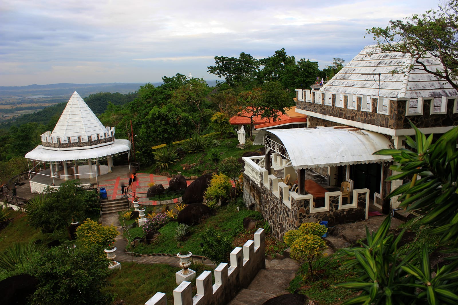 White pavilions with crenellated walls and conical roofs among gardens on a hilltop, looking out over a wide green plain under a cloudy sky.