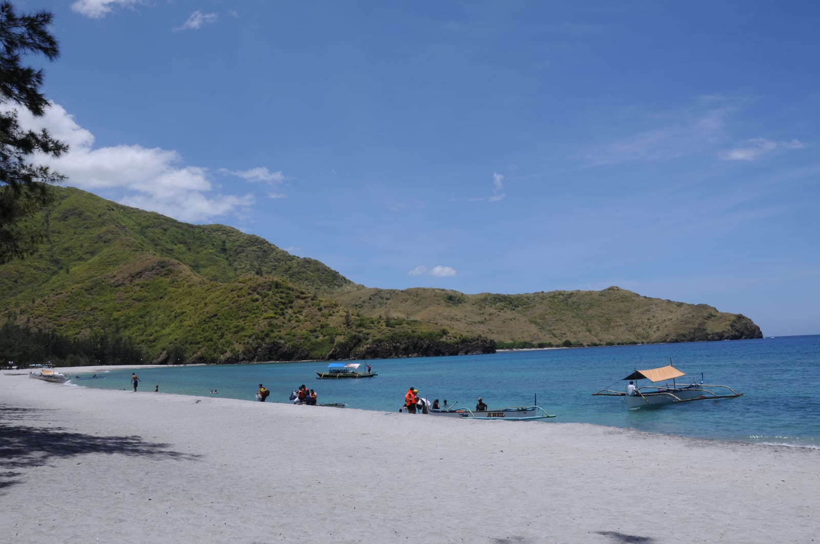 A pale-sand cove under steep green hills, with outrigger boats on clear turquoise water.