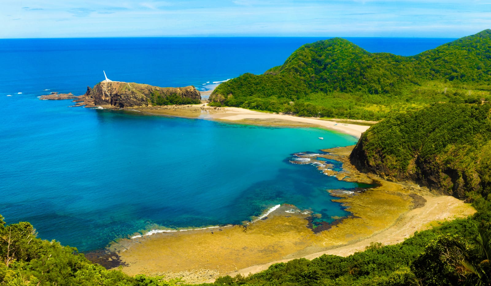 A curving bay of deep blue and turquoise water below forested hills, with a pale beach, a reef shelf, and a rocky point topped by a white lighthouse.
