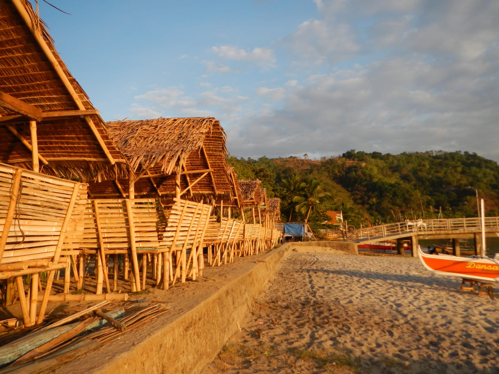 Bamboo cottages with thatched roofs along a sandy shore, an outrigger boat drawn up on the sand, and a pier at the water's edge.