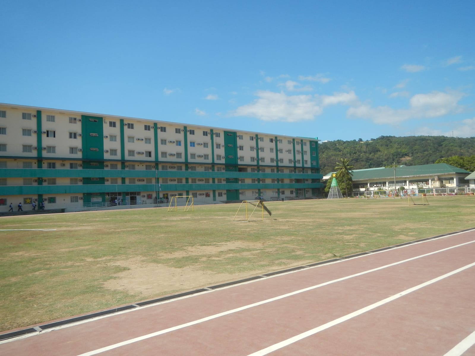 A red running track curving around a grass field, with a long multi-storey building and green hills behind.