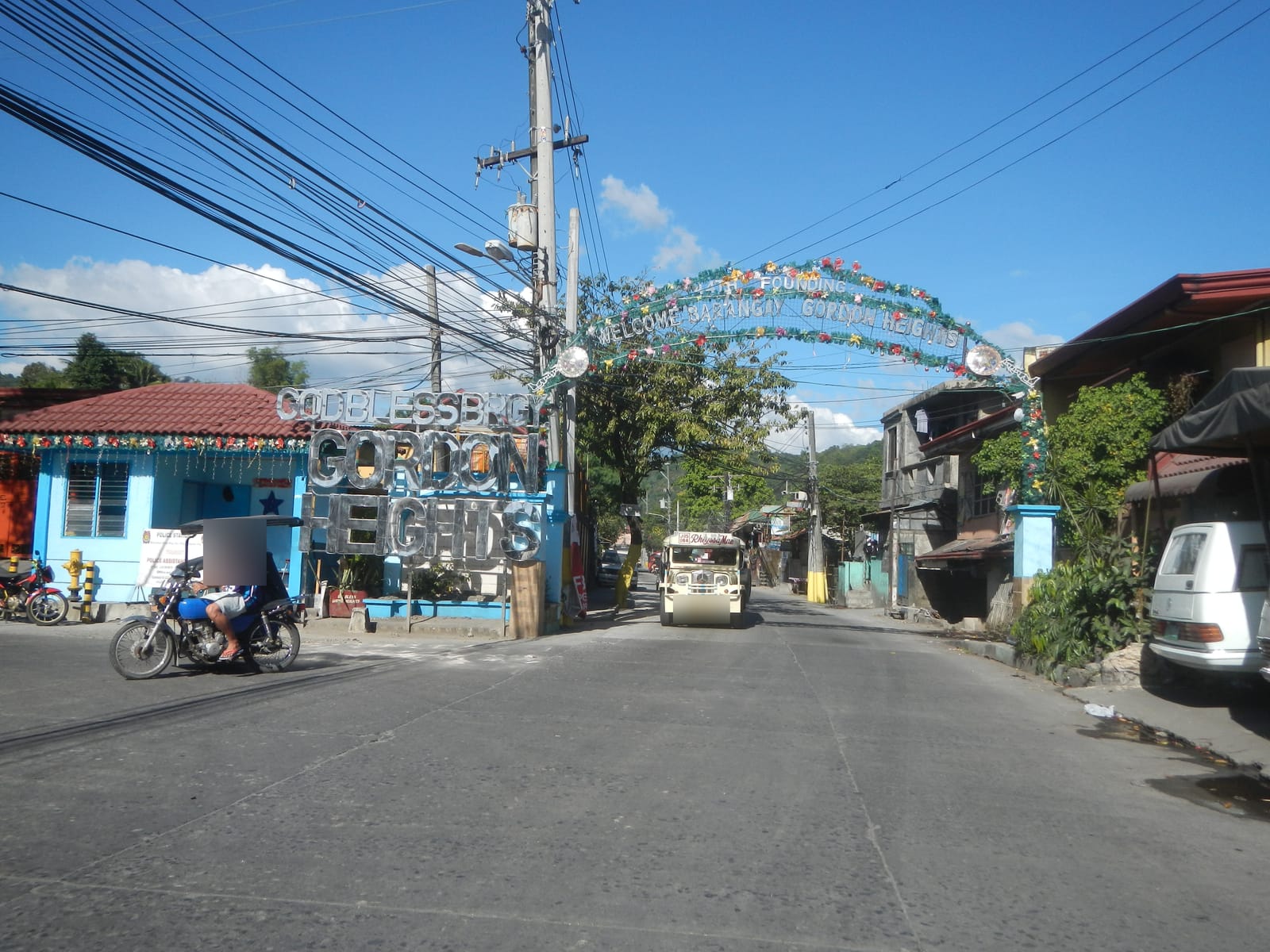 Silver letters spelling “God bless Brgy. Gordon Heights” at a street corner, with a flower-decked welcome arch and power lines above.