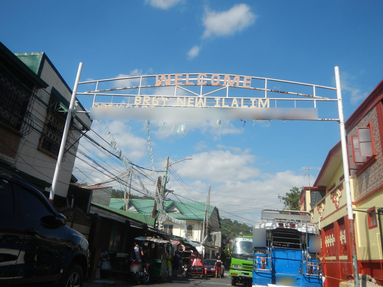 A white metal arch lettered “Welcome Brgy. New Ilalim” over a narrow street with a blue jeepney and a green truck.