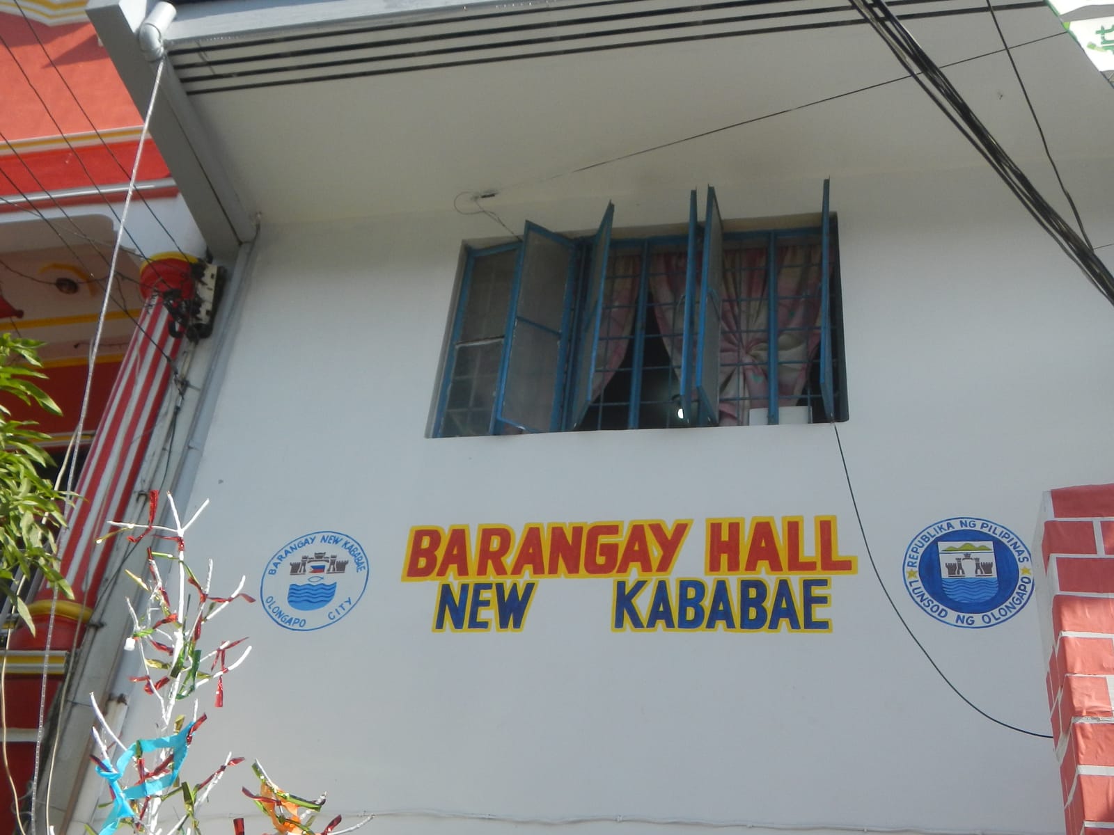 The white upper floor of a building lettered “Barangay Hall New Kababae” between two round seals, below a window with blue frames.