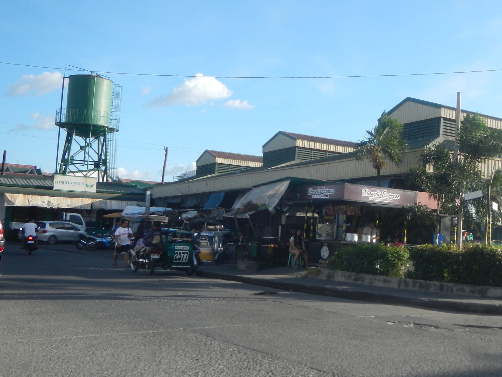 The long, low roofs of a market behind a planted traffic island, with a green water tower to the left.