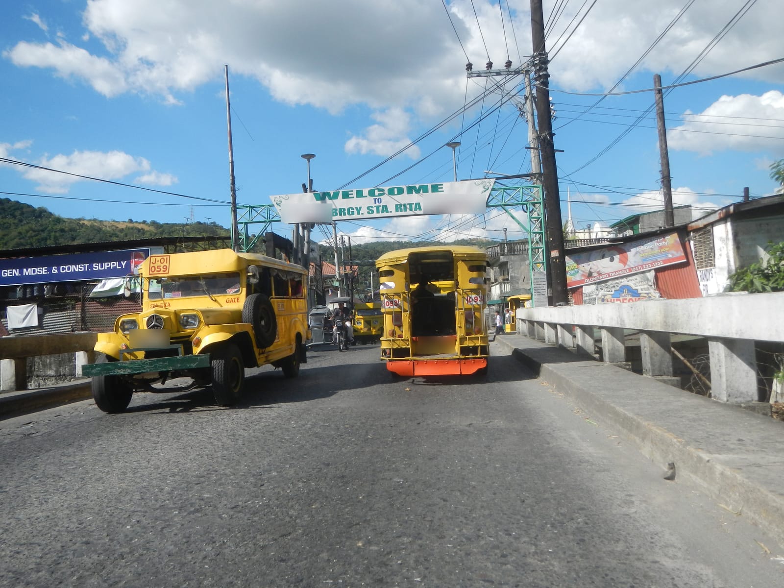 Two yellow jeepneys on a bridge under a green steel arch and a sign reading “Welcome to Brgy. Sta. Rita”, with green hills beyond.