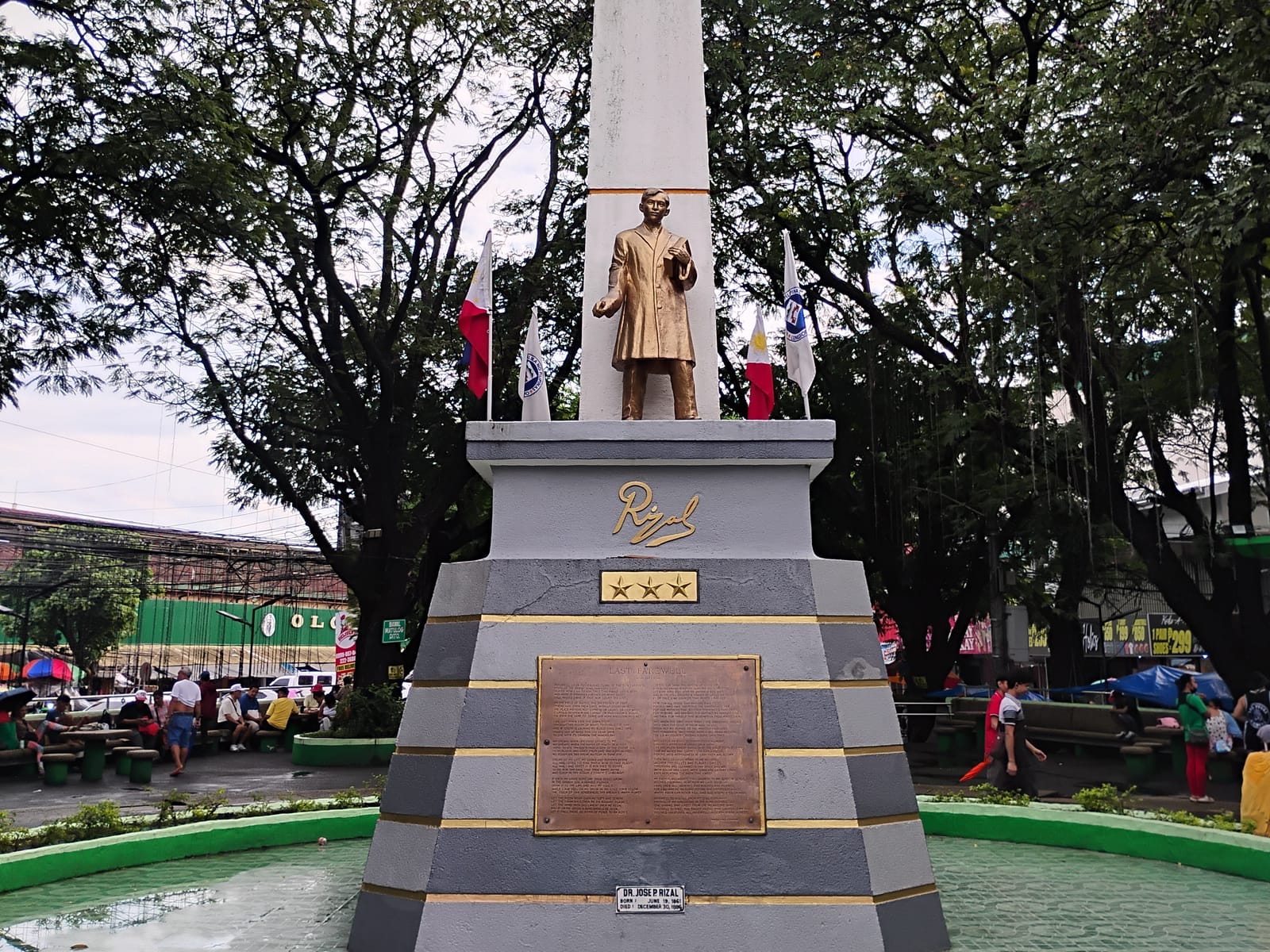A gilded statue of José Rizal on a grey stepped base before a white obelisk, between flags, under trees.
