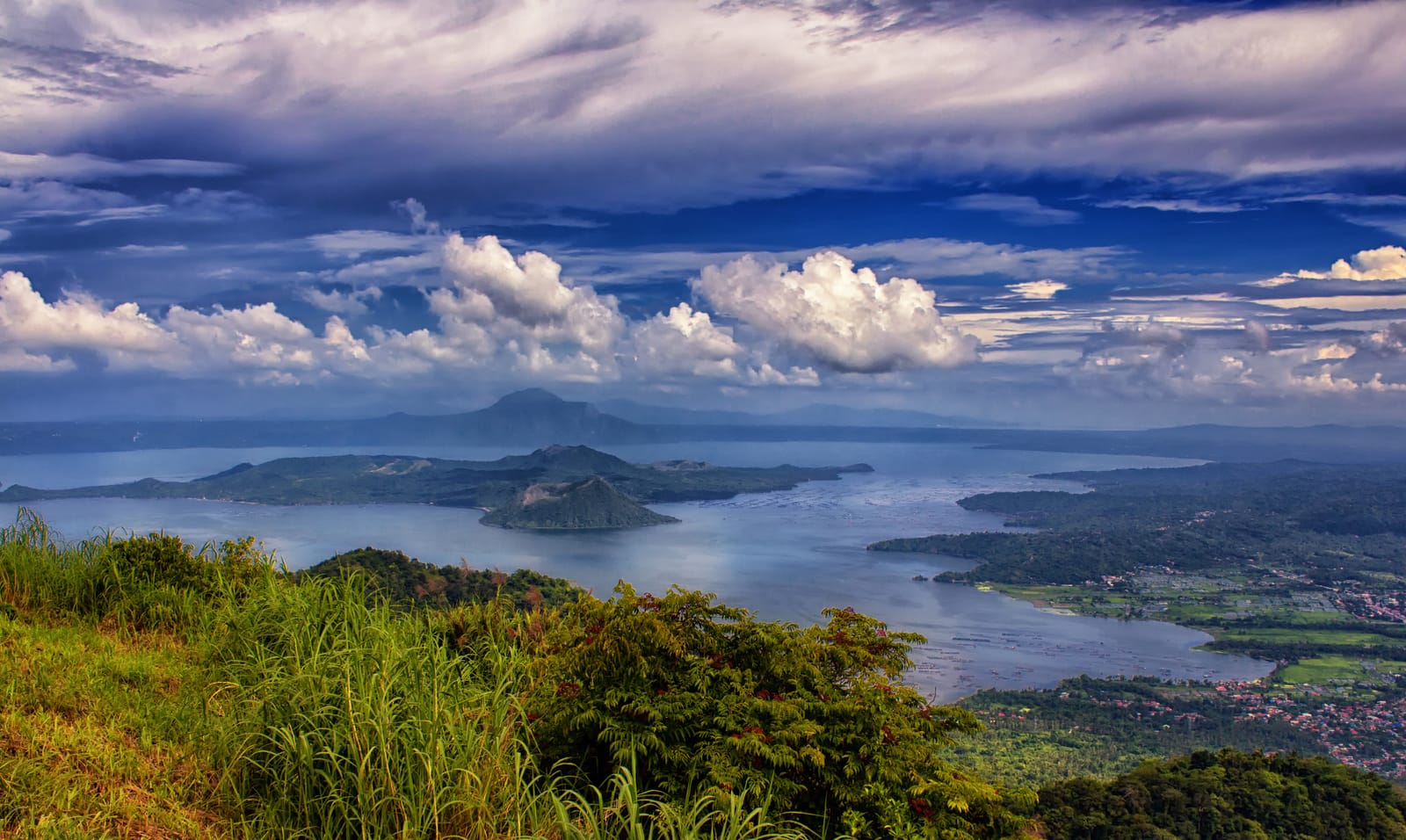 A wide blue lake with a dark volcanic island and a small cone in it, under towering white clouds, seen from a grassy ridge.