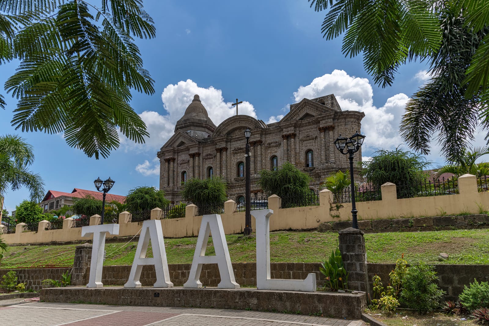 Tall white letters spelling “TAAL” on a lawn below a huge domed stone church, framed by palm fronds under a blue sky with white clouds.