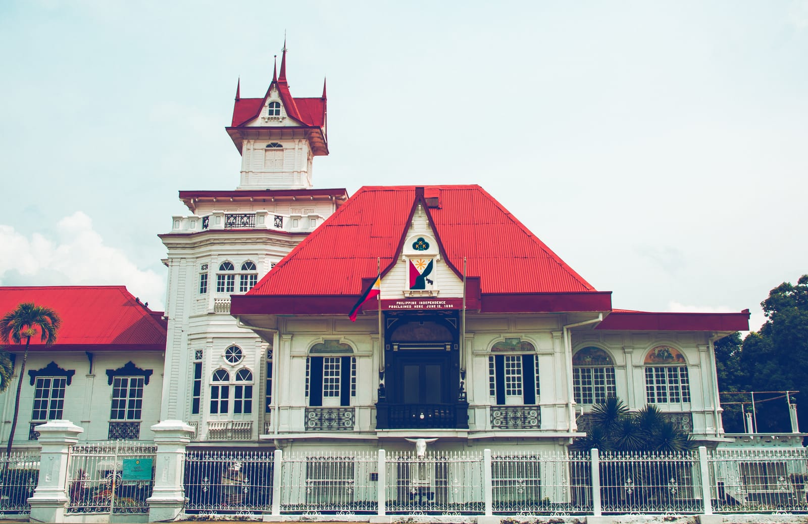 A large white mansion with steep red roofs, a balcony over the main door hung with a flag emblem, and a slender tower topped by a red spire, behind a white iron fence.