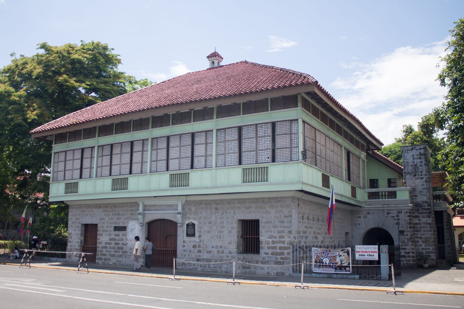A two-storey house with a stone ground floor, a pale green upper floor lined with sliding shell windows and a red-tiled roof, beside a street under a blue sky.