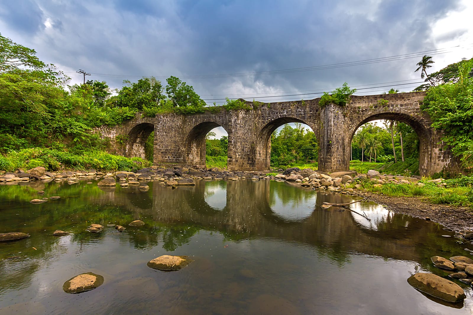A long old stone bridge of several arches over a shallow, rocky river, mirrored in the still water, under a stormy grey sky.