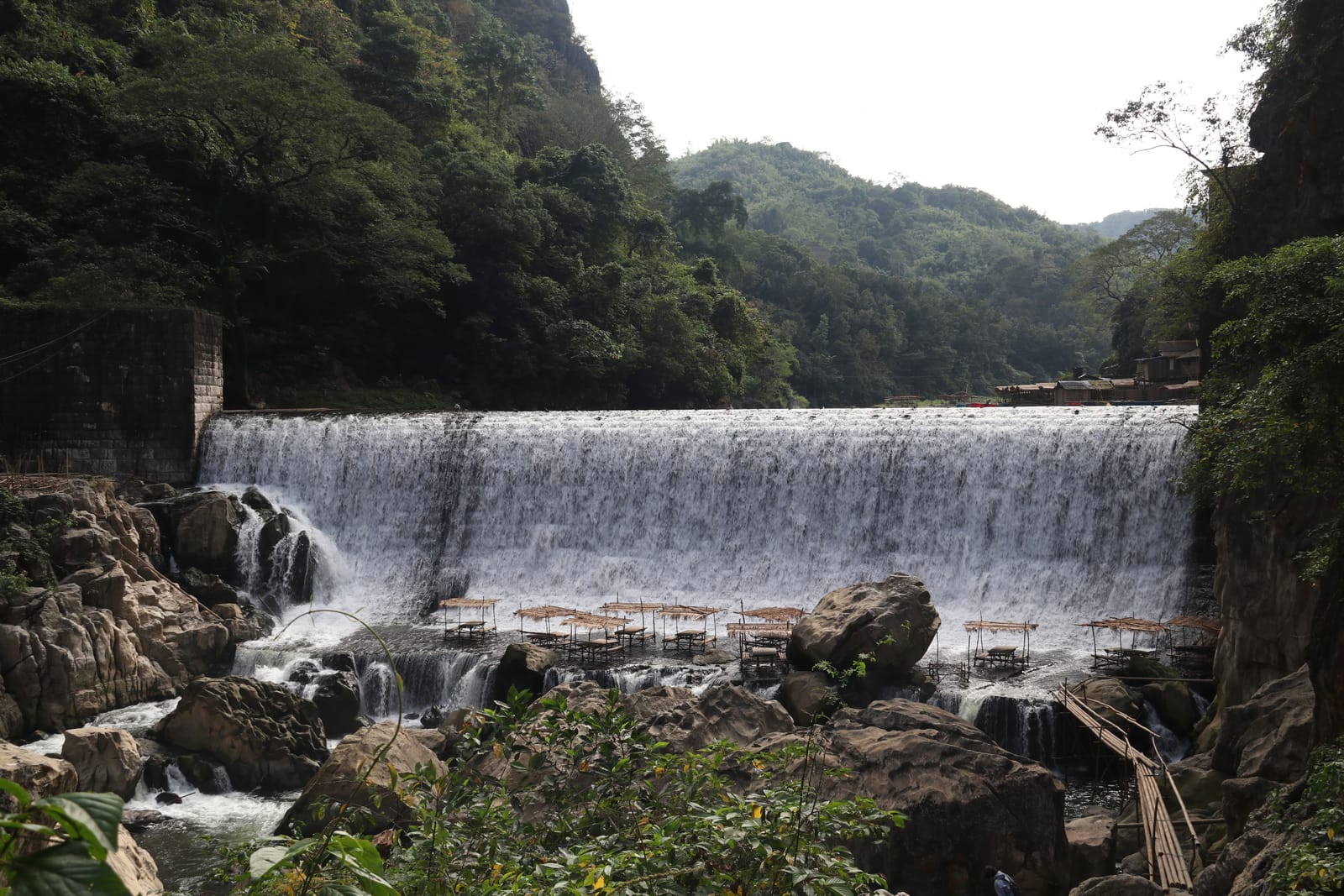 White water pouring over a long curved dam wall into a rocky river gorge, with bamboo rafts below and forested hills behind.