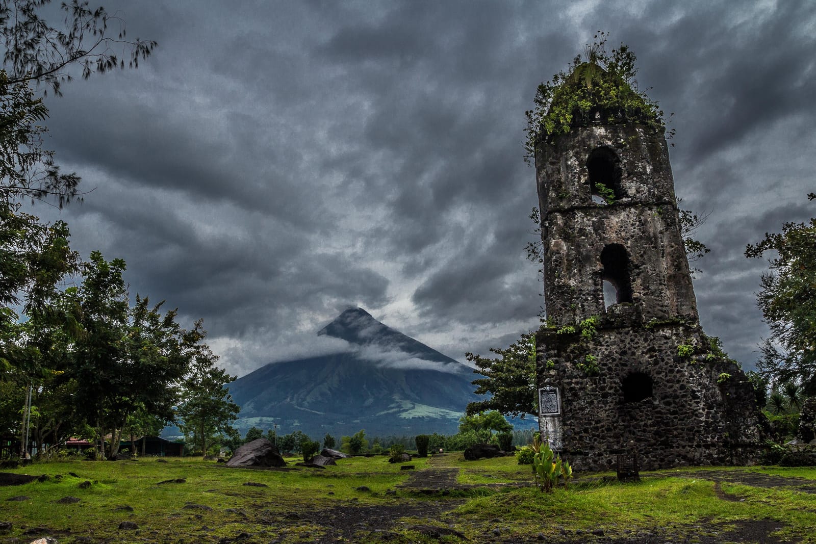 A ruined stone bell tower crowned with plants on a grassy field, with the cone of a volcano rising behind it under heavy grey clouds.