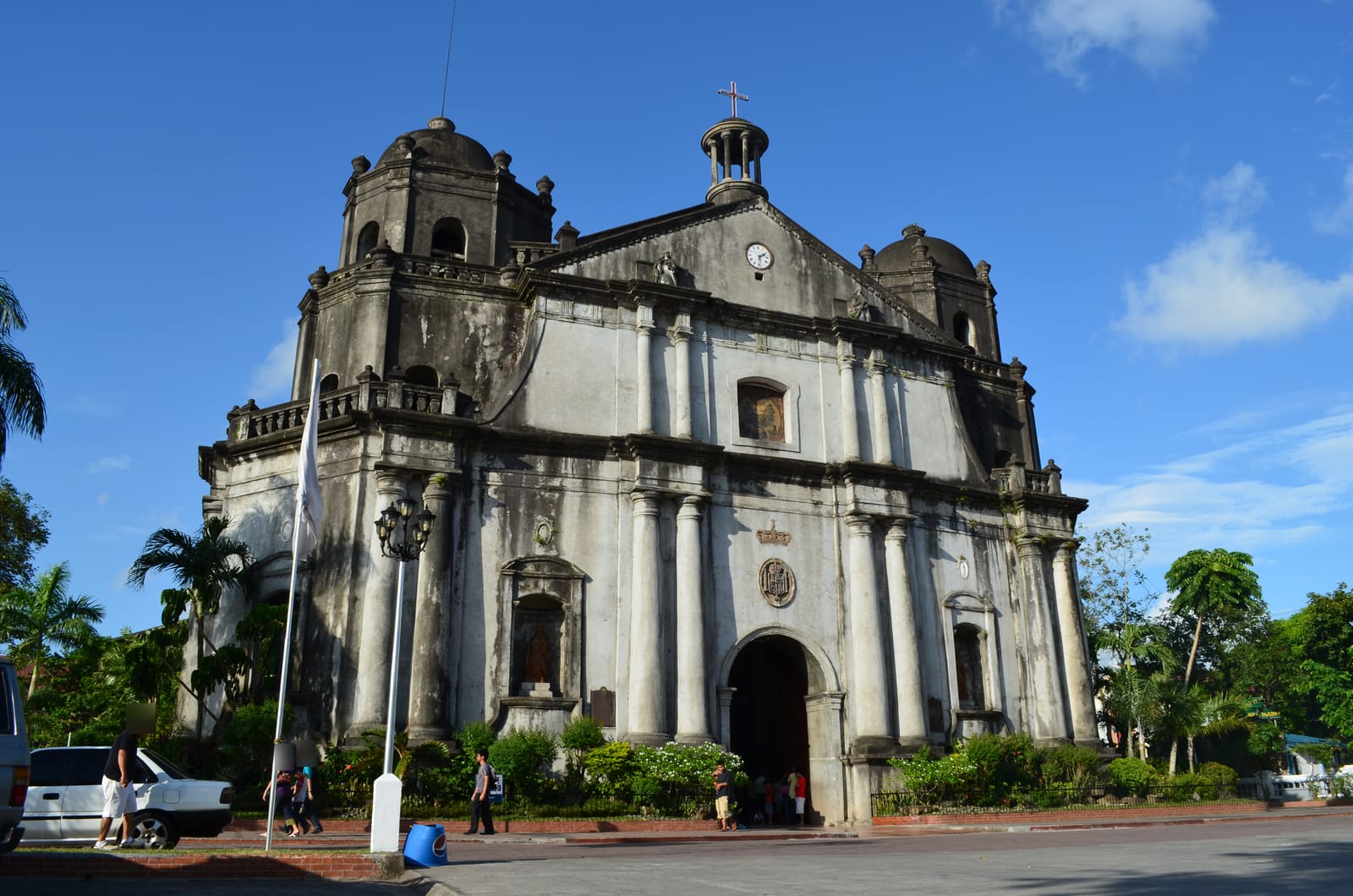 A weathered grey stone cathedral with a columned facade, a clock in its pediment and two domed bell towers, under a blue sky.