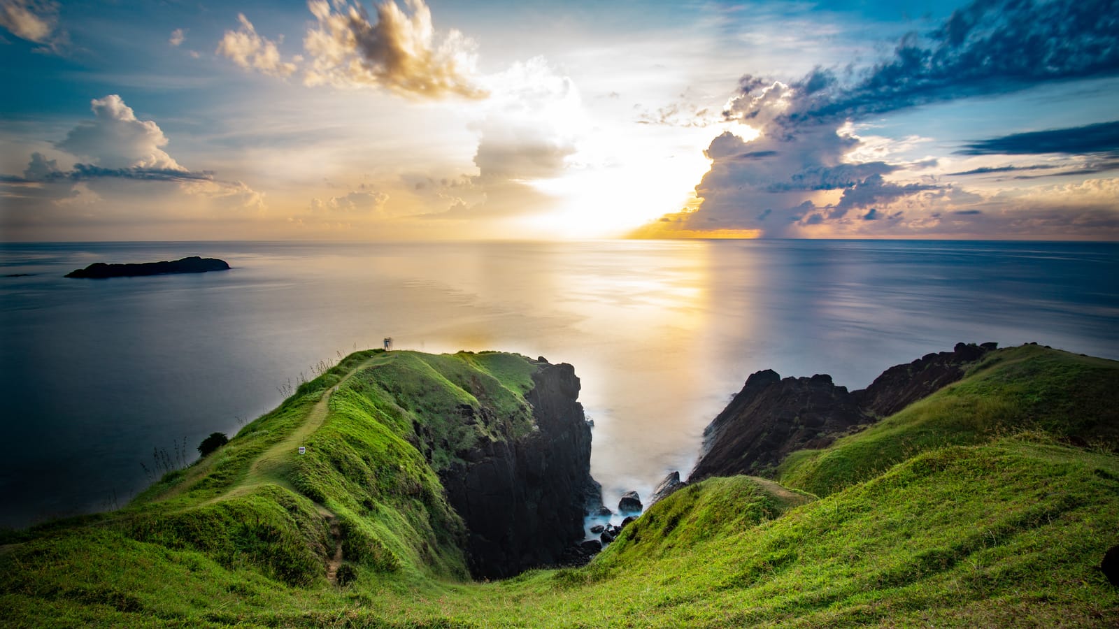 Steep green headlands and dark cliffs dropping into a calm sea, with a small island offshore and the sun breaking through clouds over the horizon.