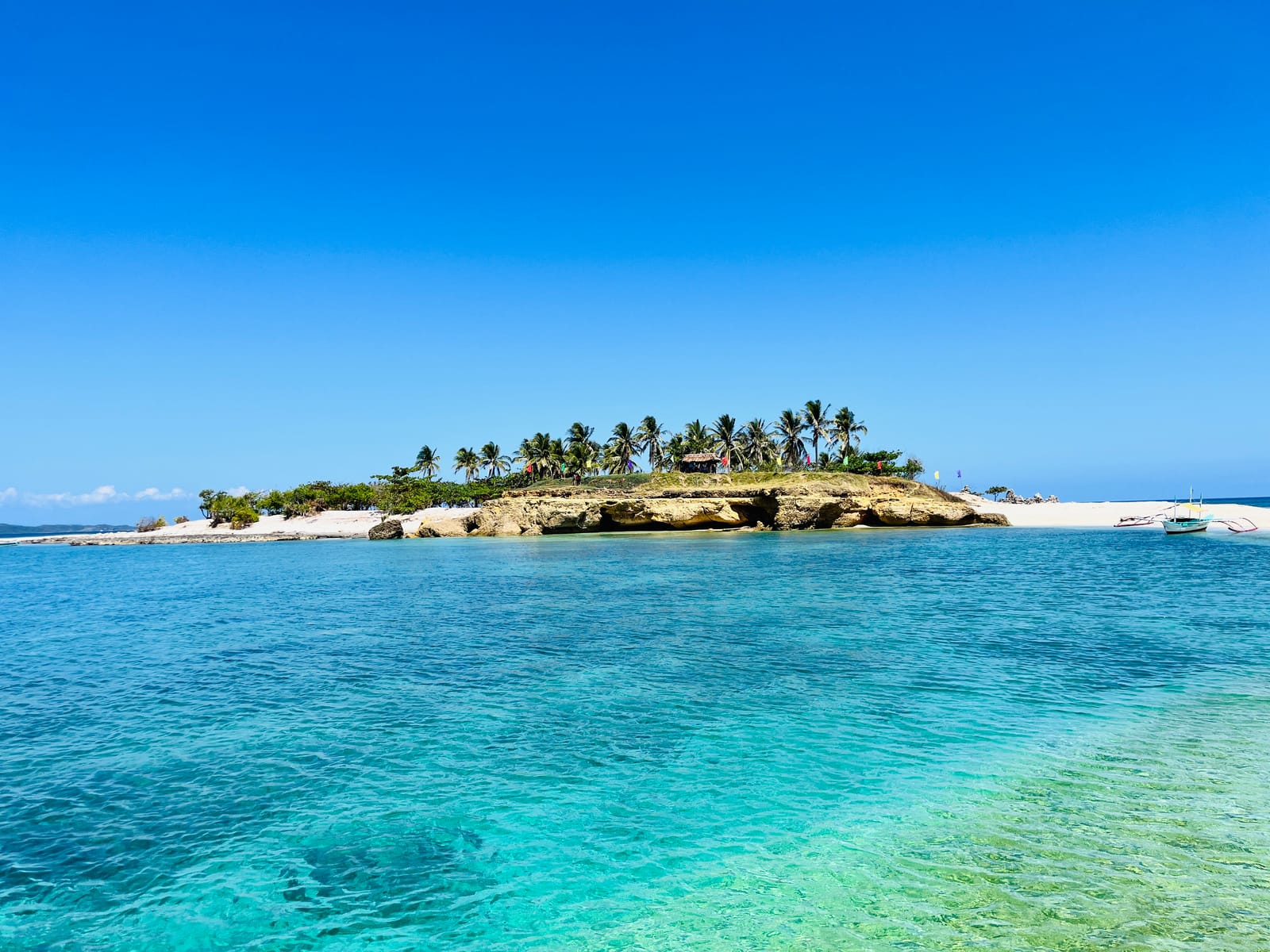A low rocky islet topped by coconut palms and a hut, ringed by white sand, across clear turquoise shallows under a cloudless blue sky.
