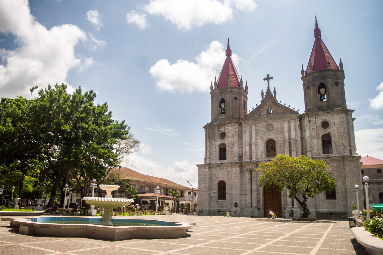 A grey stone Gothic church with two towers capped by red spires, beside a paved plaza with a white tiered fountain and shade trees, under a blue sky.
