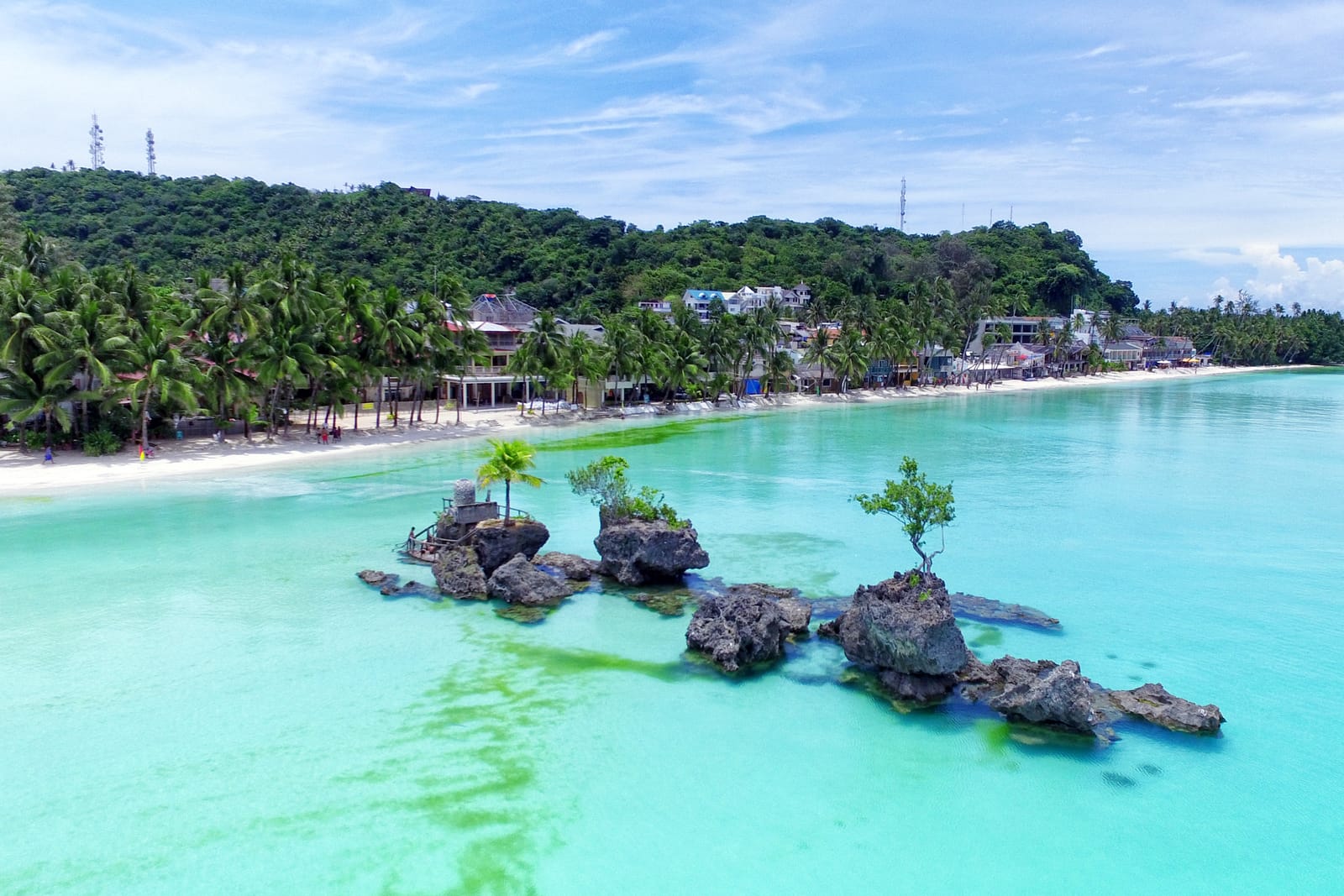A cluster of dark rocks topped by small trees and a little shrine, standing in clear turquoise shallows off a long white beach lined with palms and low resorts, below a green hill.