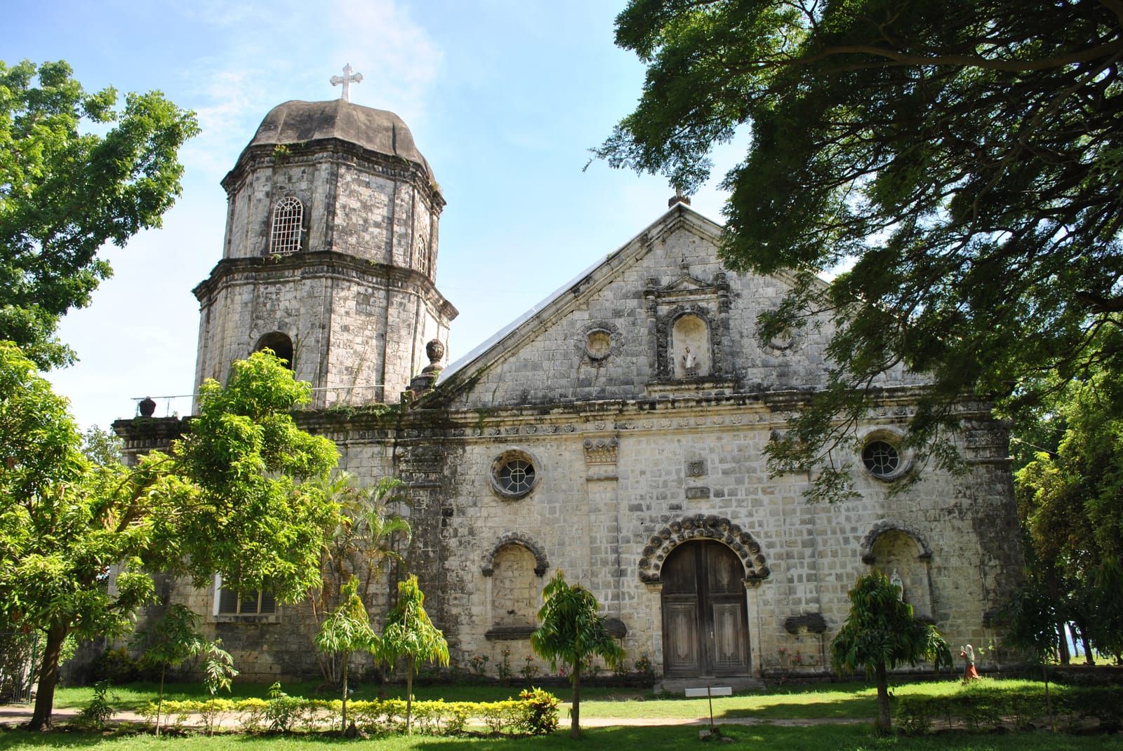 A pale coral-stone church with a carved gable and an arched wooden door, beside a squat bell tower capped by a dome and a cross, half framed by green trees under a blue sky.
