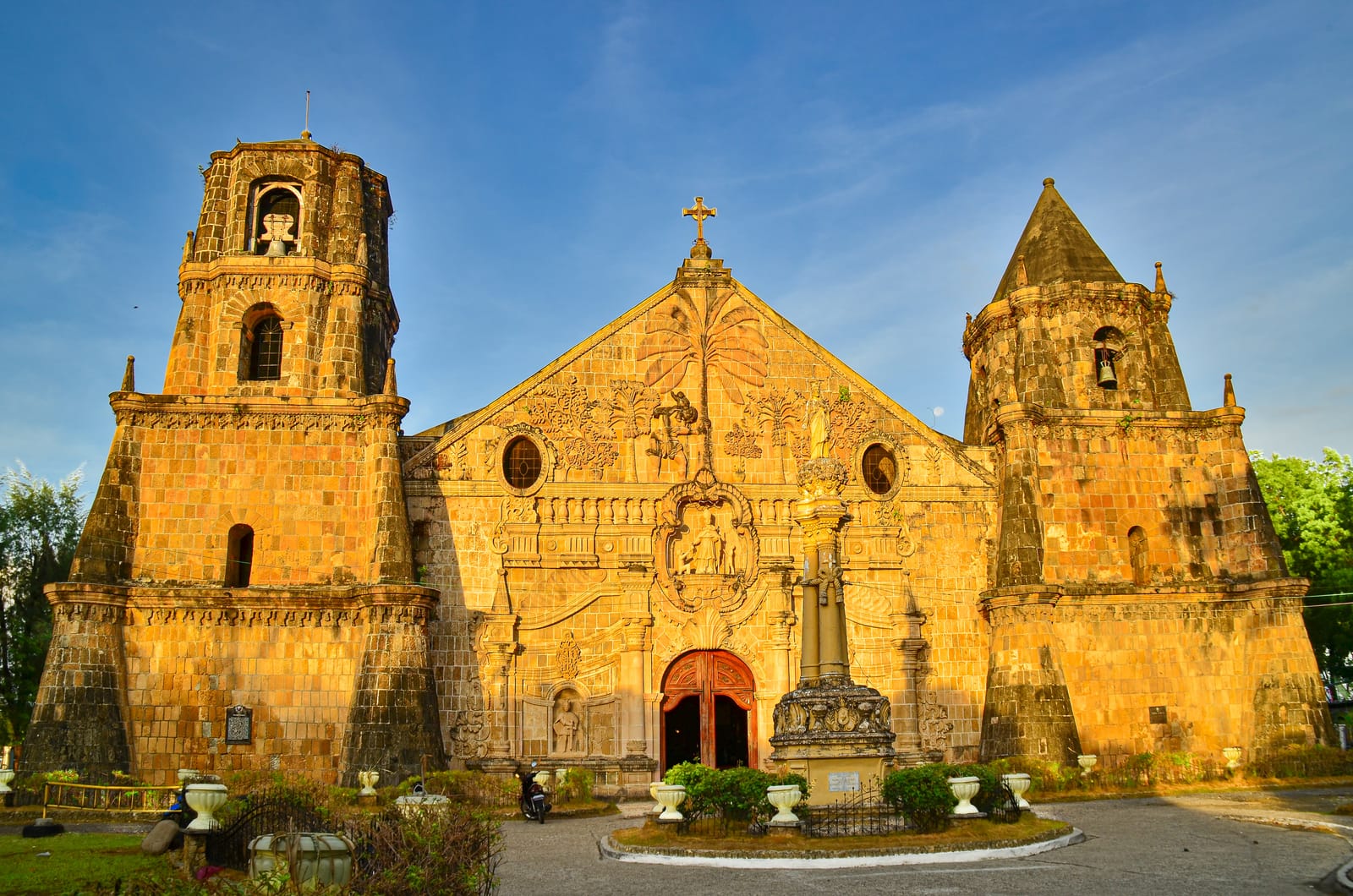 A massive stone church lit gold by the early sun, its carved gable set between two broad bell towers of unequal height, the lower one with a pointed roof, behind a small garden of white urns, under a deep blue sky.