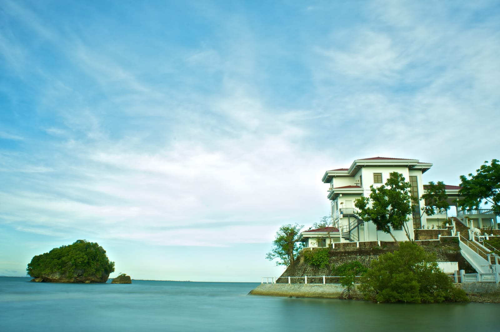 A white mansion with red roofs and wide verandas on a walled rock at the water's edge, with mangroves at its foot and a small wooded islet out in a calm sea under a pale blue sky.