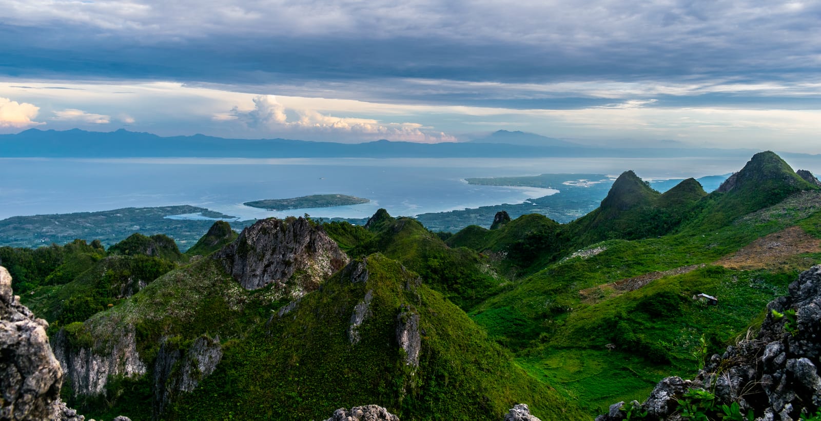 Jagged green hills and rocky peaks above a calm sea with a low island and a far coastline, under a cloudy sky.