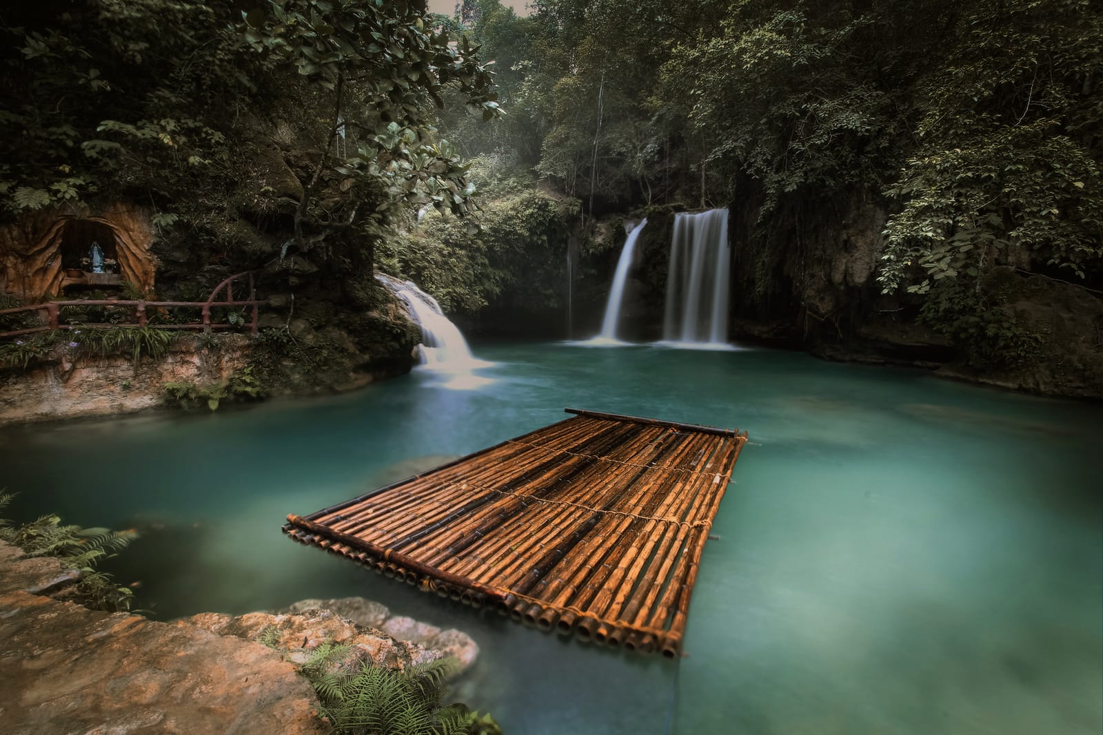 A turquoise pool below a twin waterfall in green forest, with a bamboo raft floating on the water.