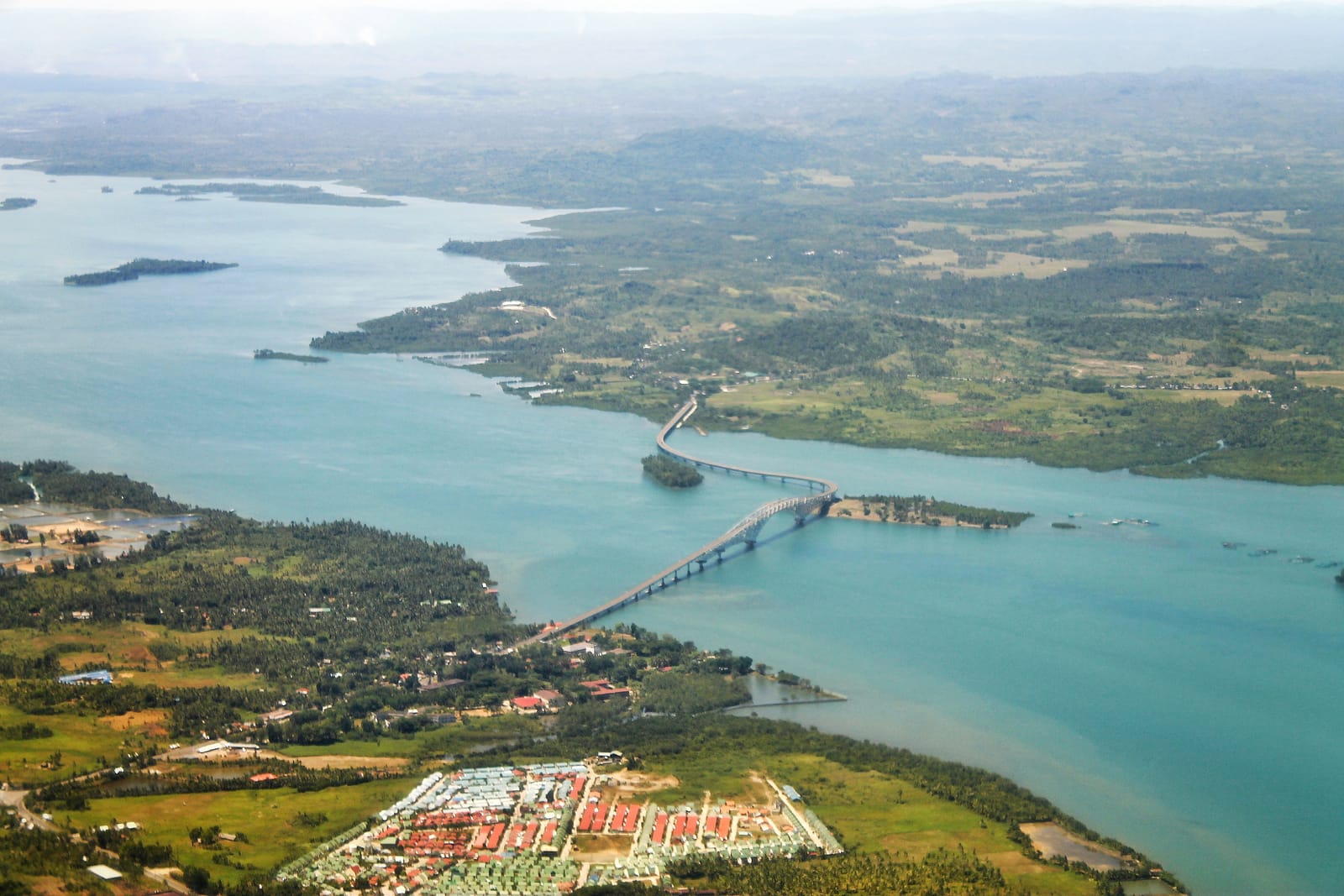 A long bridge curving across a blue strait between green shores, seen from the air, with a housing estate on the near shore and low islands beyond.