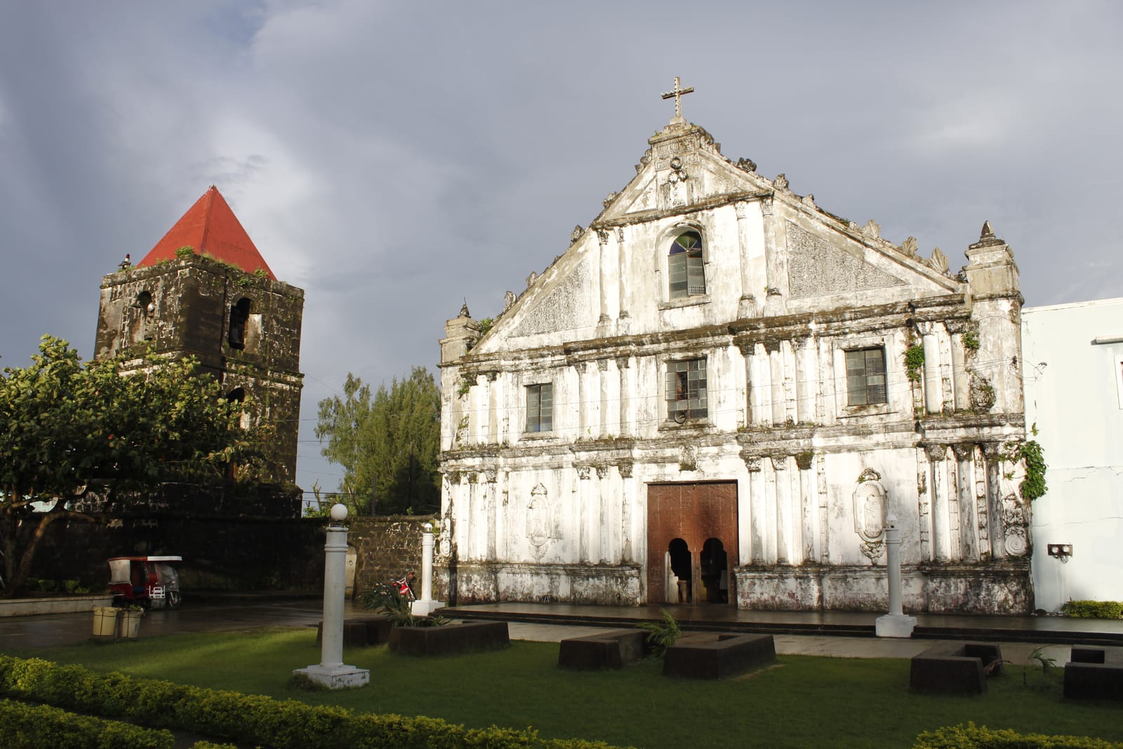 A whitewashed baroque church front with carved panels, pilasters and an arched wooden door, lit warm by a low sun against a dark storm sky, beside a stone bell tower with a red pointed roof.