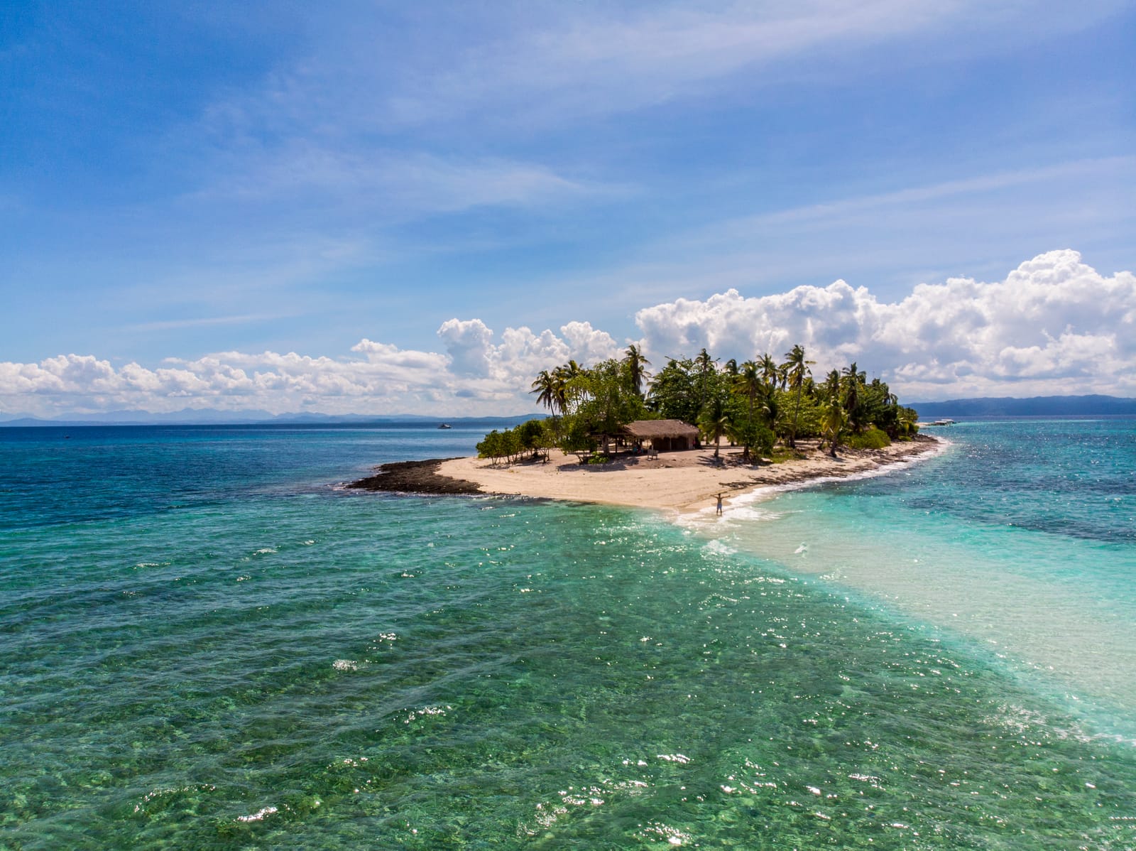 A small island of palm trees and a thatched hut, its white sandbar running out into clear turquoise shallows, seen from the air under a blue sky with white clouds.