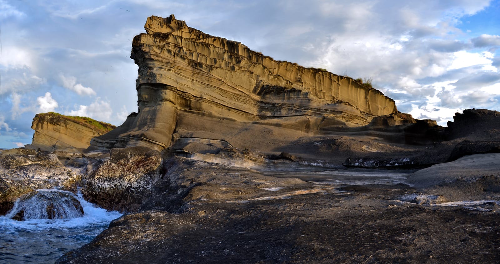 A tall, layered sandstone rock rising from a dark wave-cut shore, its stripes sloping down to the right, with surf breaking over the rocks at left under a cloudy blue sky.