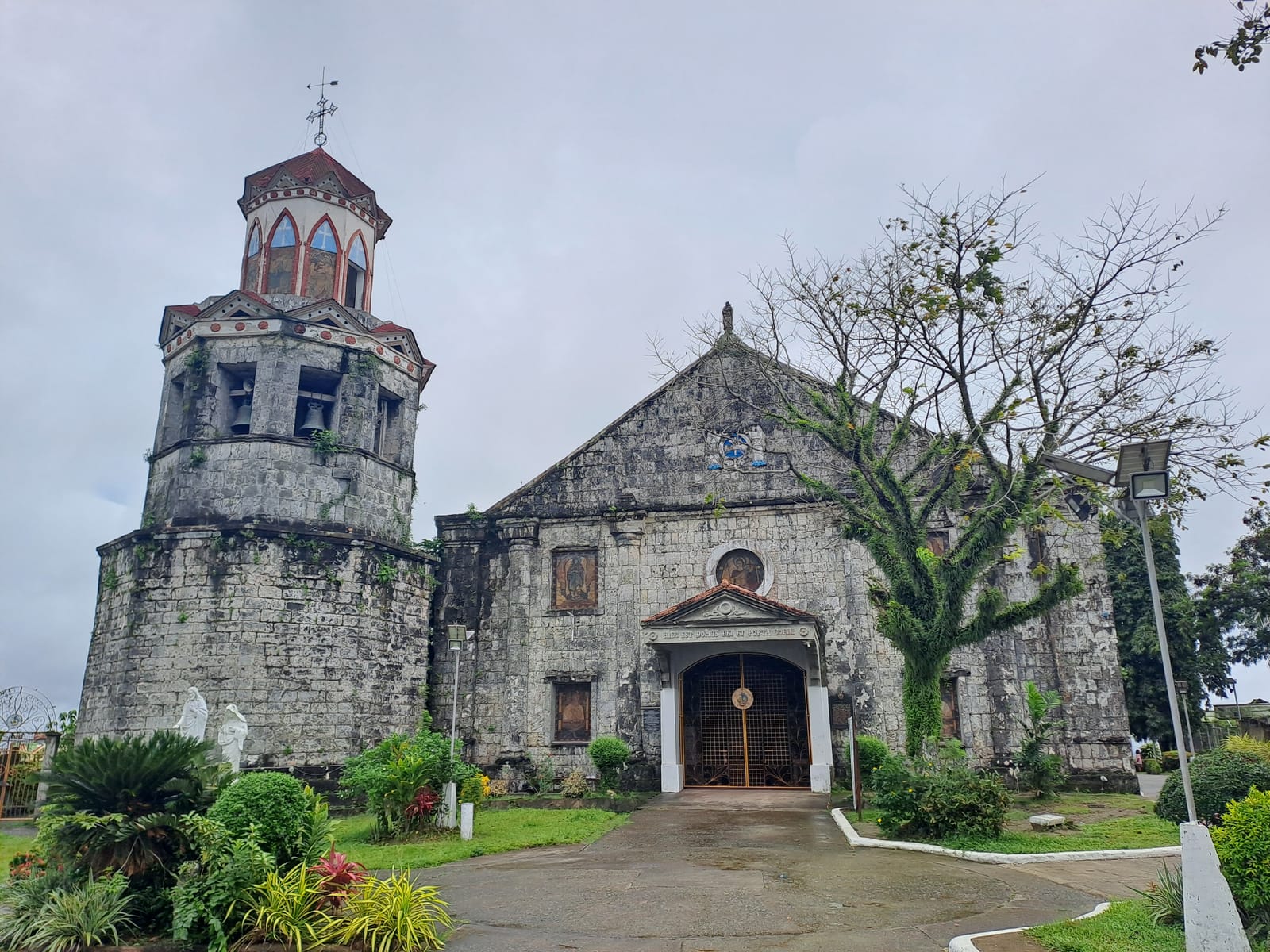 A weathered grey stone church with a pointed gable, a round window and a white-framed doorway, beside a thick round bell tower capped by a red-roofed lantern, behind a garden, under an overcast sky.