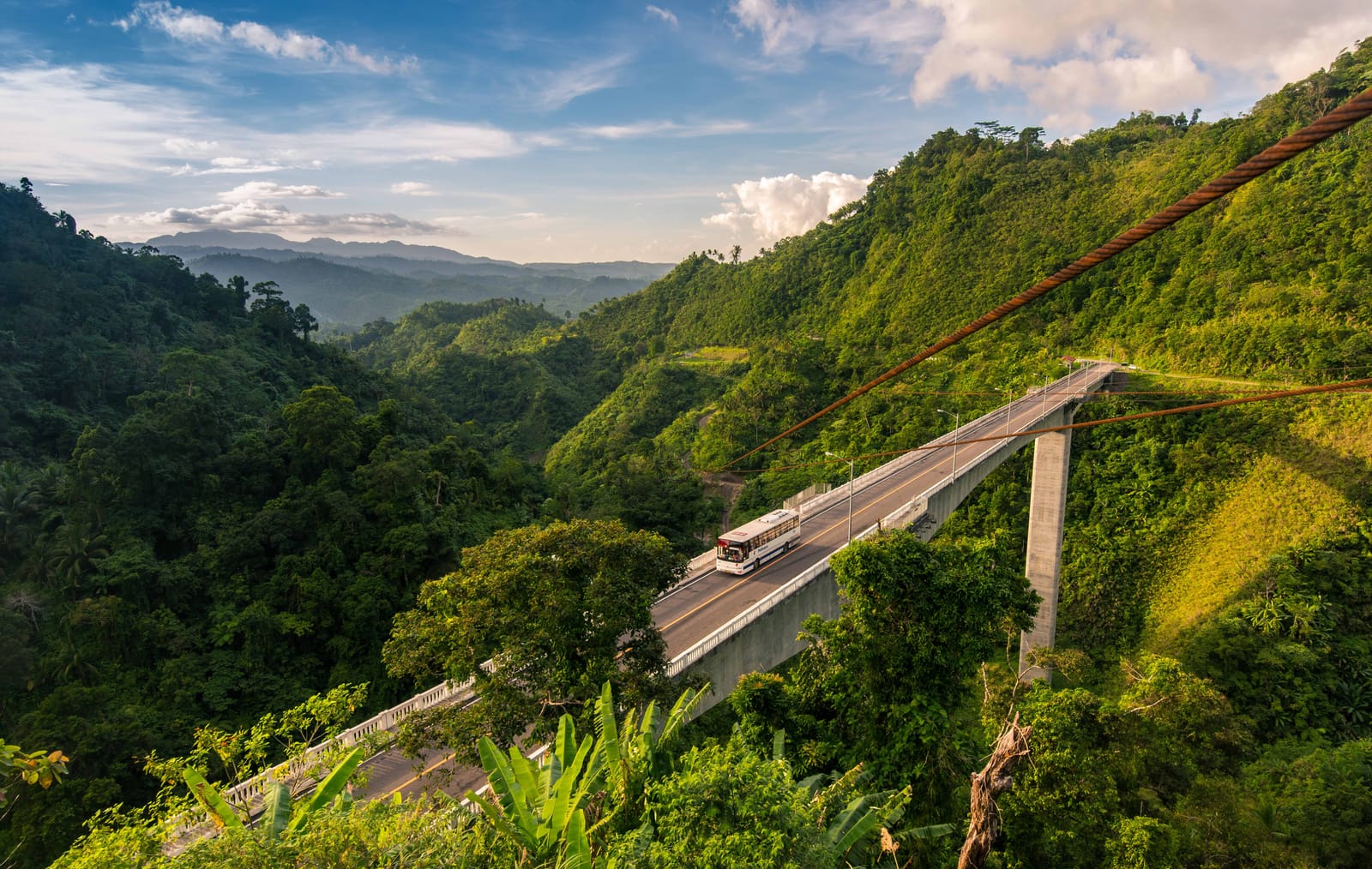 A tall concrete bridge on a single slender pier spanning a deep green valley, with a white bus crossing it and forested hills rolling away to blue mountains under a bright sky.
