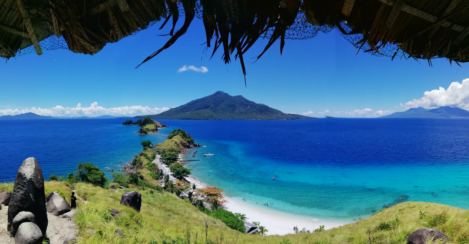 A chain of small green islets strung along a white beach and turquoise shallows, seen from a grassy hilltop under the eaves of a thatched roof, with a cone-shaped island rising from the deep blue sea beyond.