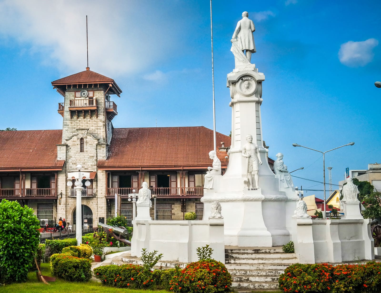 A long two-storey building with a red-tiled roof and a stone clock tower, behind a tall white monument topped by a statue and ringed by smaller statues, under a blue sky.