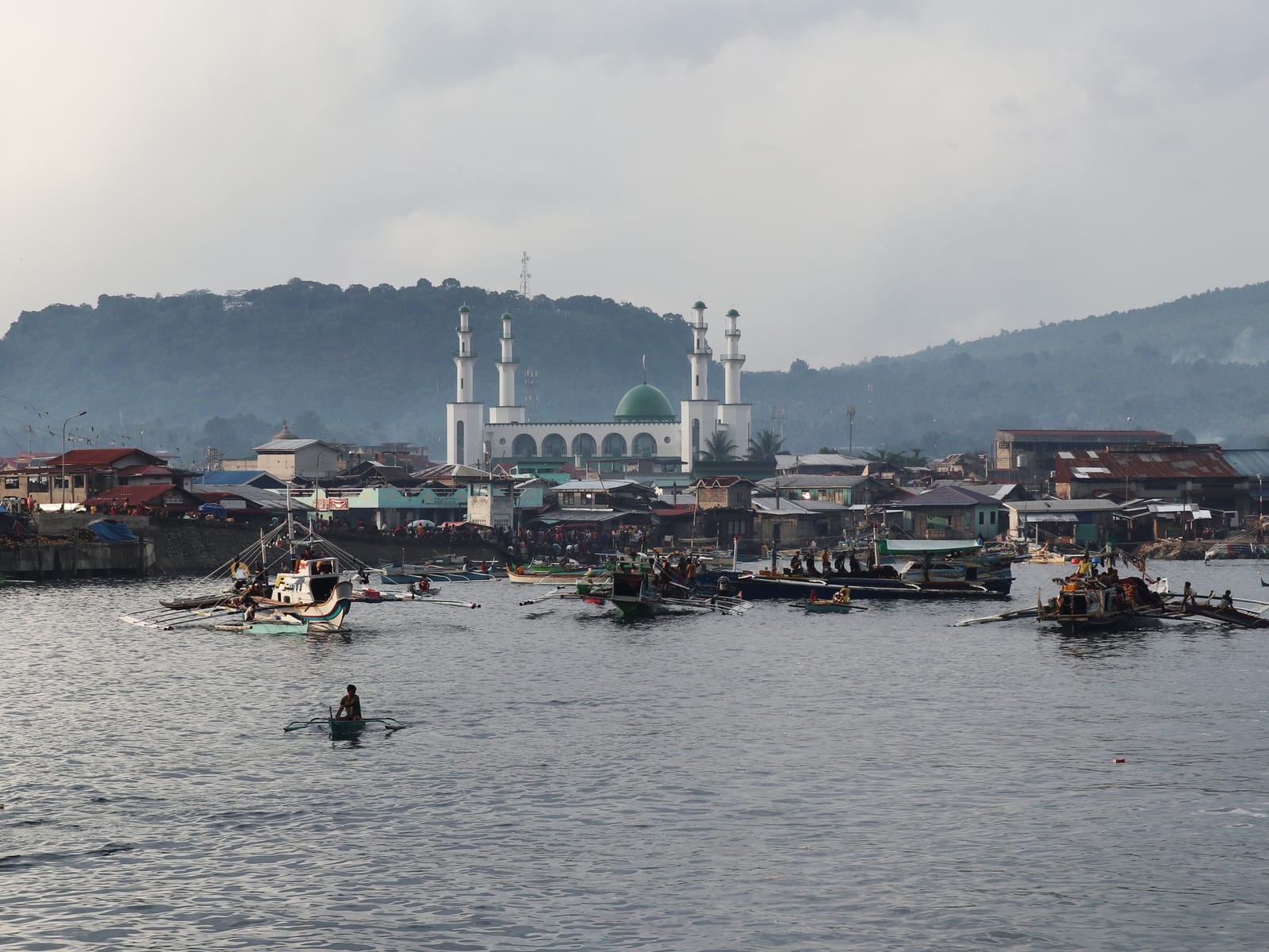A white mosque with a green dome and four slender minarets rising behind the roofs and stilt houses of a waterfront town, outrigger boats crowding the grey water in front, below a forested hill.